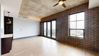 This single-floor urban loft showcases a compelling blend of industrial architecture and contemporary design. The main living area is defined by striking exposed brick walls, a raw concrete ceiling, and light hardwood flooring that together create a warm yet edgy atmosphere. A ceiling fan adds a functional accent overhead, while large black-framed French doors open directly onto a private balcony, flooding the space with natural light and offering expansive city views.

The galley-style kitchen is well-appointed with dark espresso cabinetry, stainless steel appliances including a refrigerator, range, and microwave, and a mosaic tile backsplash that adds visual texture. Light countertops provide a clean contrast to the rich cabinetry, and pendant lighting illuminates the workspace. Generous built-in storage and open shelving line one side of the kitchen, enhancing both form and function.

The bathroom is thoughtfully finished with large-format floor tiles, a dark vanity with a white countertop, and a soaking tub fully surrounded by classic white subway tile extending floor to ceiling. Chrome fixtures and wall sconces complete the polished, spa-like feel. Throughout the space, the interplay of raw industrial elements and refined modern touches creates a cohesive and distinctive urban living environment.