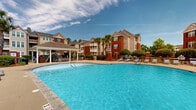 Vinings at Spanish Fort, located along US-31 in Spanish Fort, Alabama, is a thoughtfully designed residential community that combines classic Southern architectural charm with modern outdoor amenities. The community is anchored by a generously sized resort-style swimming pool with a freeform shape, fountain features, and crystal-clear water, surrounded by an expansive pool deck furnished with lounge chairs and shaded seating areas ideal for relaxation.

The clubhouse serves as the heart of the community, constructed with traditional red brick and white trim, complemented by large arched windows, white columns, and a covered brick-paved front porch furnished with classic rocking chairs. A welcoming "Vinings at Spanish Fort" entry mat greets residents at the front door, reinforcing the community's identity. The exterior patio areas extend from the clubhouse, offering additional lounge seating and open-air gathering spaces.

The grounds are meticulously landscaped with manicured shrubs, seasonal plantings, tall palm trees, and mature hardwoods, contributing to a lush, resort-like atmosphere. A covered pergola-style gazebo at the poolside provides shaded seating for outdoor dining or socializing. Volleyball court facilities are also visible within the recreation area. The multi-story residential buildings feature brick and neutral-toned exteriors with private balconies, overlooking the beautifully maintained common areas throughout the community.