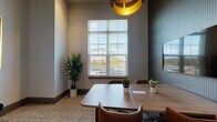 Located at 40000 Argento Way in Kannapolis, NC, this single-floor meeting space combines contemporary design with functional elegance. The conference room serves as the centerpiece, featuring a sleek wooden table surrounded by rich brown leather swivel chairs that offer both comfort and style. A large wall-mounted flat-screen display is set against a striking dark textured accent wall, providing a sharp visual contrast that enhances the room's modern aesthetic. White plantation shutters frame a generous window that fills the space with natural light and offers views of the surrounding neighborhood. A statement gold pendant light fixture hangs overhead, adding a warm, sophisticated touch to the room's atmosphere. Lush potted plants are thoughtfully placed throughout, introducing a refreshing natural element. Adjacent to the conference room, a welcoming corridor features soft neutral walls adorned with bold, gold-framed abstract artwork, complemented by a navy velvet bench with accent pillows resting on a speckled neutral carpet. This transitional area connects seamlessly to a broader common area visible in the background, hinting at additional collaborative spaces. The overall ambiance is polished, professional, and inviting, making this space well-suited for focused meetings and productive gatherings.