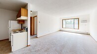 Located at 11800 Eldorado St NW in Coon Rapids, Minnesota, this one-bedroom, one-bathroom unit offers a practical and efficient single-floor layout. The open-concept living and kitchen area forms the heart of the space, featuring soft carpeted flooring, neutral white walls, and a textured ceiling. A large sliding window with a wood-trimmed frame fills the living area with natural light and provides pleasant outdoor views of surrounding greenery, while a baseboard heating system and wall-mounted air conditioning unit ensure year-round comfort. The kitchen is positioned adjacent to the living space and includes wood-tone cabinetry and essential appliances. A pendant light fixture highlights the dining area near the kitchen, adding a subtle decorative touch. The bedroom is a well-proportioned, quiet retreat with its own wood-framed window overlooking the outdoors, carpeted flooring, and baseboard heating. Two closets provide ample storage throughout the unit. The bathroom features a full tub and shower combination, a wood-vanity with an integrated sink and countertop, a large mirror, and classic mosaic tile flooring. Clean, simple, and functional, this unit offers a comfortable living environment in a residential Coon Rapids neighborhood.