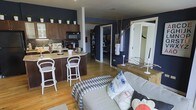This single-floor space makes a strong visual impression with its deep navy blue accent walls, which provide a striking contrast against crisp white trim and warm honey-toned hardwood flooring throughout. The open-concept layout connects the kitchen and living area in one cohesive, flowing space that feels both functional and inviting.

The kitchen is anchored by rich dark wood cabinetry topped with light-colored countertops and a curved breakfast bar with seating for two, creating a natural gathering spot. A mosaic tile backsplash adds texture, while the upper cabinet tops are styled with an array of small potted plants, bringing a touch of greenery to the space. Black appliances, including a microwave and range, complete the kitchen's modern aesthetic.

The living area is comfortably furnished and artfully decorated, featuring a neutral upholstered sofa accented with patterned throw pillows. A geometric harlequin-print area rug grounds the seating area, while eclectic decorative elements — including twisted branches, a large mirror, and sculptural accents — lend the space a curated, artistic character. A hallway beyond leads to additional areas of the home, hinting at further functional spaces. Overall, the design balances bold color choices with warm natural materials for a lively yet comfortable ambiance.