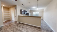 This one-bedroom, one-bathroom unit in Longview, Texas offers a practical and well-appointed layout across a single floor. The open-concept kitchen and living area serves as the heart of the home, featuring wood-look laminate flooring, neutral beige walls, and a ceiling fan that enhances airflow throughout the space. The kitchen is equipped with stainless steel appliances — including a refrigerator, electric range, and over-the-range microwave — complemented by dark-toned cabinetry and light countertops. A peninsula-style counter provides a natural division between the kitchen and living area, while a pendant light fixture adds a decorative touch above the dining space. A window allows natural light to brighten the area. Built-in open shelving along one wall offers convenient storage and display space. The bedroom is carpeted for comfort, featuring a ceiling fan, a large window with outdoor views, and a closet with double doors providing ample storage. The bathroom includes a dark wood vanity with a light countertop, a mirror, and wood-look flooring consistent with the rest of the unit. Throughout, the design is clean and functional, with consistent neutral tones creating a cohesive and move-in-ready atmosphere.