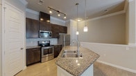 This single-story unit offers a thoughtfully designed layout with quality finishes throughout. The kitchen serves as a central highlight, featuring dark espresso cabinetry that extends to the ceiling, a subway tile backsplash, and a full suite of stainless steel appliances including a range, microwave, and refrigerator. A peninsula island anchors the space, topped with speckled granite countertops and a double stainless steel sink, illuminated by elegant pendant lighting. Track lighting overhead provides additional task illumination. Crown molding and wainscoting details add architectural refinement to the open living area, which flows seamlessly from the kitchen. Light wood-style flooring runs consistently throughout the unit, contributing to a bright, cohesive aesthetic.

The bedroom is a calm, neutral space featuring the same light flooring, crown molding, and a window fitted with horizontal blinds that allows natural light while maintaining privacy. The bathroom connects to a walk-in closet, providing practical storage. Dark wood vanity cabinetry with granite countertops ties the bathroom's finishes back to the kitchen, maintaining a consistent design language throughout the unit.