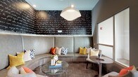This thoughtfully designed single-floor lounge space blends contemporary aesthetics with comfortable, functional furnishings. The interior is anchored by a standout curved banquette upholstered in soft grey, adorned with an eclectic mix of patterned and jewel-toned throw pillows in mustard yellow, burnt orange, and navy. Above it, a dramatic black accent wall features flowing white script text, adding an artistic, expressive focal point to the seating nook. A large dome pendant light illuminates the area, complementing the warm, inviting atmosphere.

Adjacent to the lounge seating is a sleek kitchenette bar area, complete with a light stone countertop, stainless steel refrigerator, wall-mounted flat-screen television, and a row of colorful mixed-metal stools. Orange butterfly wall art adds a playful, artistic touch overhead. Tile flooring with geometric-patterned area rugs grounds each zone with visual structure.

The entry area features a bold large-scale abstract painting alongside a bistro-style table and modern lattice chairs, creating an artful welcome. Natural light filters in through large windows, while a balcony offers sweeping views of a well-maintained green field and modern urban buildings beyond, connecting the interior to its dynamic surroundings.