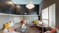 This thoughtfully designed single-floor lounge space blends contemporary aesthetics with comfortable, functional furnishings. The interior is anchored by a standout curved banquette upholstered in soft grey, adorned with an eclectic mix of patterned and jewel-toned throw pillows in mustard yellow, burnt orange, and navy. Above it, a dramatic black accent wall features flowing white script text, adding an artistic, expressive focal point to the seating nook. A large dome pendant light illuminates the area, complementing the warm, inviting atmosphere.

Adjacent to the lounge seating is a sleek kitchenette bar area, complete with a light stone countertop, stainless steel refrigerator, wall-mounted flat-screen television, and a row of colorful mixed-metal stools. Orange butterfly wall art adds a playful, artistic touch overhead. Tile flooring with geometric-patterned area rugs grounds each zone with visual structure.

The entry area features a bold large-scale abstract painting alongside a bistro-style table and modern lattice chairs, creating an artful welcome. Natural light filters in through large windows, while a balcony offers sweeping views of a well-maintained green field and modern urban buildings beyond, connecting the interior to its dynamic surroundings.