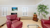 This single-floor townhome offers a thoughtfully designed open-concept layout that seamlessly connects the living, dining, and kitchen areas. The living room is a standout space, featuring soaring vaulted ceilings that amplify the sense of openness, light hardwood flooring, and vertical panel wall accents that add architectural character. Elegant French doors with frosted glass panes serve as a refined focal point, providing both visual interest and connectivity between spaces.

The dining area transitions naturally from the living room, distinguished by neutral carpet flooring and a raised dining table with accompanying bar-height chairs. Adjacent to the dining space, a recessed kitchen nook is equipped with warm wood cabinetry, black appliances including a refrigerator and microwave, and a breakfast bar with seating — a compact yet functional cooking space.

A separate carpeted area extends toward what appears to be a stairwell landing, suggesting additional access points within the unit. Multiple windows fitted with roller shades allow for adjustable natural light throughout. The overall palette of warm beige tones, white trim, and crown molding creates a cohesive, welcoming ambiance throughout the space.