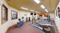 Located at 1617 E Main Ave in Puyallup, WA, this well-equipped fitness center offers a comprehensive workout environment across a single floor. The space is thoughtfully organized to accommodate a variety of exercise needs, from cardiovascular training to strength building.

The main workout area is bright and open, featuring recessed tray ceiling details and a grid of flush-mounted fluorescent panel lights that ensure consistent, even illumination throughout. Warm yellow-toned walls and red-hued carpet contribute to an energizing yet inviting atmosphere. Multiple flat-screen televisions are mounted on the walls, providing entertainment for those using the cardio equipment.

Cardio equipment is well-represented, with a row of Life Fitness treadmills, an elliptical trainer, and a recumbent stationary bike, all positioned on protective rubber mats. A full dumbbell rack with a wide range of free weights serves those focused on resistance training, while a dedicated room houses a multi-station cable and weight machine along with a weight bench, ideal for targeted strength exercises. Windows with warm-toned blinds allow natural light into this area and offer views of the surrounding greenery.

The facility also includes an accessible restroom, sanitation dispensers, and a clearly posted fitness room notice. The layout provides a comfortable, functional space suitable for a variety of fitness routines.