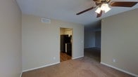 This single-floor unit features an open layout that connects the main living area to additional rooms and a kitchen. The living space is finished with wall-to-wall carpet in a neutral tone, complemented by beige walls and white baseboards that create a clean, understated aesthetic. A ceiling fan with an integrated light fixture is mounted to the textured ceiling, providing both airflow and ambient lighting. An HVAC vent is visible near the upper wall, ensuring climate control throughout the space. A doorway from the main living area leads directly into the kitchen, which contrasts with hardwood-style flooring. The kitchen is equipped with black appliances, including a refrigerator and a range, alongside white cabinetry offering ample storage. A secondary opening from the living area leads to an additional room, also carpeted, suggesting a bedroom or flexible-use space. The overall layout is practical and straightforward, with a functional flow between areas. The neutral color palette and consistent finishes throughout give the space a cohesive and move-in-ready appearance.