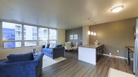 This single-floor space features an open-concept layout that seamlessly connects the kitchen, dining, and living areas, creating a cohesive and functional environment. Rich, dark hardwood flooring extends throughout, providing a warm and grounding contrast to the lighter wall tones. The living area is anchored by a large area rug and benefits from generous natural light streaming through floor-to-ceiling windows along one wall, offering views of the surrounding exterior. A charcoal accent wall with framed artwork adds depth and visual interest to the dining zone, where a round table with upholstered chairs creates an intimate setting. Pendant lighting with frosted globe shades hangs above the kitchen peninsula, which features dark cabinetry and a neutral countertop surface. The peninsula serves as both a functional workspace and a natural visual divider between the kitchen and the living space without disrupting the open flow. A textured stone or tile column detail adds an architectural element between the living and kitchen zones. Overall, the space presents a contemporary aesthetic with a balanced mix of neutral tones, natural materials, and modern lighting.