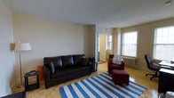 This single-floor space centers around a well-appointed living room that balances comfort and functionality. The room features warm parquet hardwood flooring throughout, lending a classic and timeless character to the interior. Neutral-toned walls provide a clean backdrop, while the generous ceiling height adds to the sense of openness.

Natural light flows in abundantly through a large, multi-pane window on one side of the room, complemented by a classic cast-iron radiator beneath it — a nod to the building's traditional construction. A dark leather sofa anchors the seating area, paired with a deep red armchair and matching ottoman, creating a comfortable and inviting arrangement. A blue and white striped area rug defines the central living zone, adding a casual, relaxed visual element.

A doorway visible from the living room leads into an adjacent space, suggesting a connected layout that allows for a natural flow between rooms. The overall ambiance is warm and livable, combining traditional architectural details with practical, everyday furnishings suited for comfortable daily living.