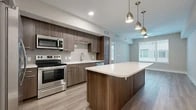 This well-designed one-bedroom, one-bathroom unit offers a thoughtfully arranged floor plan that maximizes its compact footprint. The open-concept kitchen and living area serves as the heart of the space, anchored by a substantial kitchen island with a crisp white quartz countertop and warm wood-toned cabinetry. The kitchen is fully equipped with a complete suite of stainless steel appliances, including a French door refrigerator, range, built-in microwave, and dishwasher. A geometric hexagonal tile backsplash adds a stylish focal point, while light hardwood-style flooring runs consistently throughout, unifying the space. Pendant lighting above the island complements recessed ceiling fixtures, providing layered illumination. A ceiling fan serves the living area, which benefits from natural light through large windows fitted with blinds. The bedroom is a comfortable size, featuring the same hardwood-style flooring, a ceiling fan, and a window that brings in natural light. A dedicated closet provides ample storage. The bathroom features a wide wood-toned vanity with a white countertop, a large mirror, tile flooring, and a full tub-shower combination with classic white subway tile surround.
