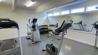 This fitness room offers a clean, functional environment designed to accommodate a range of exercise routines. The space features a light-toned, neutral color palette with white walls and a durable epoxy-style flooring that is both practical and easy to maintain. A row of high, horizontal windows lines the upper portion of one wall, allowing natural light to filter into the room while maintaining privacy.

The room is equipped with several key pieces of cardio equipment, including an elliptical trainer, a stationary upright bike, and an additional cardio machine, all positioned to allow comfortable use. A dedicated free weights area features a multi-tiered dumbbell rack stocked with a broad range of weights, catering to various strength training needs.

A large floor-to-ceiling mirror spans one wall, visually expanding the space and allowing users to monitor their form during workouts. The overall layout is thoughtfully organized to maximize usable floor space while keeping equipment accessible. The bright, minimal aesthetic contributes to a focused and energizing atmosphere well-suited for fitness activities.