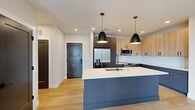 This thoughtfully designed one-bedroom, one-bathroom residence in Richmond, Virginia offers a clean, contemporary aesthetic with high-quality finishes at every turn. The open-concept kitchen and living area serves as the heart of the home, anchored by a striking blue kitchen island with a crisp white countertop and an integrated sink, illuminated by a pair of sleek black pendant lights with gold-toned hardware. The kitchen features two-tone cabinetry — warm wood upper cabinets paired with slate-blue lower cabinets accented by gold pulls — complemented by a classic white subway tile backsplash and a full suite of stainless steel appliances. Light hardwood flooring flows throughout the main living areas, enhancing the bright, airy feel. The living area is bathed in natural light from a glass exterior door and adjacent windows. The bedroom offers plush carpeting, a ceiling fan, and a window with outdoor views, while a dedicated walk-in closet provides ample built-in shelving and hanging storage. The bathroom continues the modern design language with slate-blue cabinetry, gold drawer pulls, a white countertop vanity, a tub-shower combo, and matte black fixtures, all tied together with light wood-look flooring.