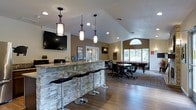 This single-story entertainment lounge is designed for social gatherings and recreation, combining stylish finishes with a comfortable, open layout. The space is anchored by a prominent bar counter finished with a stacked stone facade and a granite-style surface, accompanied by sleek black adjustable bar stools with chrome bases. Three decorative pendant lights hang above the bar area, adding warmth and visual interest to the vaulted ceiling, which rises toward an arched window at the far end of the room, inviting generous natural light throughout the space.

The flooring transitions from rich hardwood planks near the bar to a textured area rug beneath the billiards zone, defining separate functional areas within the open floor plan. A wall-mounted flat-screen television is positioned above the bar, making the space well-suited for watching sports or entertainment. Recessed lighting supplements the pendant fixtures, ensuring consistent illumination throughout.

Toward the rear of the space, a full-size pool table is surrounded by casual seating, creating a dedicated game area. The warm neutral wall tones, combined with decorative framed artwork and thoughtful lighting, give the lounge an inviting and well-appointed ambiance ideal for leisure and social interaction.