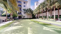 This property is set within a vibrant, resort-style community that showcases a striking blend of tropical landscaping and well-appointed outdoor amenities. The centerpiece of the exterior is a manicured putting green, offering a recreational feature rarely found in residential settings. Flagged holes dot the smooth turf, inviting both casual and enthusiastic golfers to practice their short game in an open-air setting.

The courtyard is framed by tall, mature palm trees that lend a distinctly tropical ambiance, complemented by lush hedging and ornamental plantings throughout. A paved walkway winds along the perimeter, connecting various areas of the outdoor space and offering easy access to the surrounding building entrances.

The multi-story building that borders the courtyard features balconies overlooking the green, providing residents with scenic views of the landscaped grounds. The light-colored exterior façade reflects the sunny, warm-weather character of the location.

The overall atmosphere is relaxed and resort-like, combining recreational convenience with natural beauty. The abundant greenery, open sky, and well-kept grounds create an inviting outdoor environment suited to an active and leisurely lifestyle.