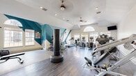 Located at 7550 S Blackhawk St in Englewood, Colorado, this fitness center offers a thoughtfully designed exercise space across a single floor. The room is defined by soaring vaulted ceilings, dual ceiling fans, and an abundance of natural light streaming through large windows, including a striking arched transom window that serves as a focal point. The interior is further elevated by bold teal geometric wall murals that lend the space a modern, energetic aesthetic.

The cardio zone features multiple treadmills, elliptical machines, and a stationary bike, all positioned to face the windows and a wall-mounted television for an engaging workout experience. The strength training area is well-appointed with a multi-station cable machine, a freestanding heavy punching bag, a dumbbell rack, kettlebells of varying weights, a weight bench, and medicine balls. A dedicated functional training section includes a TRX suspension system and stability balls, set on a smooth concrete floor ideal for dynamic movement. Full-length mirrors along one wall enhance the sense of space and allow for form checks during exercise. Durable hardwood-style flooring runs throughout the main workout areas, tying together a clean, professional environment suited for a comprehensive fitness routine.