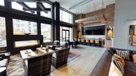 This single-floor communal space is a masterclass in modern industrial design, seamlessly blending raw materials with refined finishes. Soaring double-height ceilings and expansive floor-to-ceiling windows flood the interior with natural light, offering open views of the surrounding urban streetscape. The generous open-plan layout is thoughtfully divided into distinct functional zones, each contributing to an inviting and versatile atmosphere.

The lounge area anchors one side of the space, furnished with richly upholstered leather and plaid accent chairs arranged around a central coffee table, anchored by a patterned area rug. A sleek linear fireplace adds warmth and ambiance to this seating zone. Reclaimed wood accent elements and exposed brick walls lend an industrial character that is balanced by the warmth of light hardwood flooring throughout.

Transitioning toward the back, a full-length bar and kitchen area commands attention with dark cabinetry, pendant lighting suspended from above, and bar seating for a crowd. Multiple wall-mounted flat-screen televisions make this an ideal entertainment hub. A dining table with seating sits between the two zones, further enhancing the space's versatility for both casual gatherings and more structured events. Overall, the space exudes a sophisticated yet approachable character.