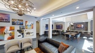 This clubhouse is a thoughtfully designed, single-floor communal space that blends contemporary aesthetics with a welcoming, social atmosphere. The open-concept layout seamlessly connects multiple functional zones, including a relaxed lounge area, a conversational seating nook, and a dining space, allowing residents and guests to move freely throughout.

The lounge area is anchored by a rich leather sofa accented with colorful throw pillows and complemented by a plush accent chair, centered beneath a striking sculptural chandelier with a swirling metallic design that serves as a dramatic focal point. Polished concrete floors run throughout, adding an industrial-chic quality to the space.

Bold, large-scale artwork adorns the walls, lending a gallery-like energy and vibrant pops of color across the interior. Structural columns define the transition between areas without interrupting the open flow. The dining area features a natural wood table paired with modern chairs, illuminated by a sleek pendant light overhead.

Recessed lighting and a tray ceiling detail enhance the architectural depth of the space, while neutral gray walls provide a sophisticated backdrop that allows the colorful art and furnishings to take center stage. Overall, the clubhouse offers an engaging, well-appointed environment ideal for socializing and community connection.