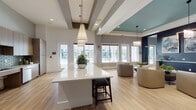This single-floor space is a thoughtfully designed amenity and community hub that blends contemporary aesthetics with functional versatility. The open-plan layout is anchored by a large white quartz island topped with elegant pendant lighting, serving as a central gathering point within the main lounge area. Light wood-tone flooring extends throughout, unifying the various zones and contributing to a warm, welcoming atmosphere.

The kitchen area is equipped with full-height wood-grain cabinetry, a teal glass subway tile backsplash, stainless steel appliances including a refrigerator and dishwasher, and generous counter space — designed to support events and social gatherings. Exposed ceiling beams and elevated ceiling heights add architectural character to the main area, while large windows and glass doors invite abundant natural light and views of the surrounding exterior.

Distinct gathering rooms are partitioned by floor-to-ceiling steel-framed glass walls, maintaining visual connectivity while providing acoustic separation. One room features a bold black-and-white striped tile accent wall alongside café-style seating, while another showcases a nature-inspired mural, dual flat-screen televisions, and patterned carpeting. A teal ceiling detail and modern drum pendant fixtures add color and design continuity across zones.