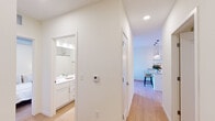 This thoughtfully designed single-floor residence offers a clean, contemporary aesthetic throughout, characterized by crisp white walls, light hardwood flooring, and an abundance of natural light. The open-concept living area serves as the heart of the home, where a large sliding glass door leads to a private balcony offering sweeping views of the surrounding cityscape. Exposed metallic ductwork adds an industrial accent to the ceiling, lending the space a modern, urban character. The kitchen is efficiently laid out with white cabinetry, stainless steel appliances including a full-size refrigerator, and a peninsula with bar seating — ideal for casual dining. A hallway connects the living spaces to the private quarters, with a thermostat panel conveniently positioned along the corridor. The bedroom is serene and well-lit, featuring balcony access and the same distinctive exposed ductwork detail. A spacious walk-in closet with wire shelving systems provides generous storage. The bathroom is clean and functional, featuring a white vanity with ample drawer storage, a wide mirror, and a shower-tub combination. A versatile flex room offers additional space suitable for a home office or study. Throughout, the residence maintains a cohesive, minimalist palette that feels both modern and welcoming.