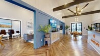Located at 3415 US-41 in Warner Robins, Georgia, this single-story leasing and community clubhouse for Grand Life offers a thoughtfully designed, coastal-inspired interior that immediately sets a welcoming tone. Upon entry, visitors are greeted by a striking reception desk adorned with the branded "Welcome to the Grand Life" signage and an illuminated logo, all set beneath soaring vaulted ceilings accented by bold dark wood exposed beams. The floors throughout feature a distinctive herringbone-patterned hardwood, adding visual texture and elegance to the open layout.

A blue shiplap accent wall serves as a central design focal point, housing a wall-mounted flat-screen TV and flanked by decorative cabinetry and lush potted plants. The lounge area provides comfortable seating alongside natural light streaming in through large windows and glass doors that offer views of the surrounding property. A dedicated kitchenette area is outfitted with white cabinetry, dark countertops, a beverage cooler, stainless steel appliances, and a dramatic herringbone-tiled backsplash with open shelving. A separate dining nook with a round wood table, cane-back chairs, and sheer-draped floor-to-ceiling windows rounds out the space. Globe chandeliers and recessed lighting complete the warm, resort-inspired ambiance throughout.