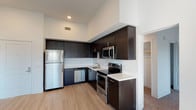 This single-floor unit offers a thoughtfully designed layout that balances modern finishes with functional living spaces. The kitchen serves as a central highlight, featuring dark espresso cabinetry that extends to the ceiling, crisp white countertops, and a full suite of stainless steel appliances including a refrigerator, electric range, built-in microwave, and dishwasher. Light wood-style flooring runs throughout the kitchen and entry areas, lending warmth and a contemporary feel. The elevated ceiling height in the kitchen area adds a sense of openness and volume to the space. The bedrooms are carpeted for comfort and benefit from natural light through window blinds, with tall ceilings that enhance the airy atmosphere. Each bedroom includes closet storage, with at least one featuring a dedicated walk-in or reach-in closet with built-in shelving and hanging rods. The bathroom is finished in a clean, minimalist style with light wood-look flooring, white tiled walls, a soaking tub with shower combination, and a dark-toned vanity with a white countertop. The overall design palette — neutral whites, warm taupes, and dark cabinetry accents — creates a cohesive and modern aesthetic throughout the unit.