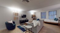 This well-appointed single-floor studio offers a seamless open-concept layout that efficiently combines living, sleeping, dining, and cooking areas into one cohesive space. Light hardwood flooring runs throughout, lending warmth and continuity across every zone.

The living area serves as the central hub, furnished with a plush gray sofa, a navy accent chair, and a marble-topped side table. A wall-mounted flat-screen TV anchors one side, complemented by a floating dark media console and abstract artwork that adds a contemporary artistic touch. A stylish area rug defines the lounge zone.

The sleeping area occupies one end of the studio, benefiting from two large double-hung windows that invite natural light. A platform bed with teal accent pillows is paired with a wooden dresser and a compact study desk with a leaning shelf, making the space functional for both rest and work.

The kitchen is fully equipped with warm wood cabinetry, granite countertops, a white subway tile backsplash, and stainless steel appliances including a refrigerator, dishwasher, and range. A compact dining table with leather chairs sits just adjacent, creating a natural eat-in area.

The bathroom features tile flooring, a wood-toned vanity, and a full bathtub with shower combination, finished in a clean, neutral palette throughout.