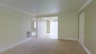 This single-story space presents a clean, open layout bathed in natural light. The main living area features wall-to-wall carpet in a neutral beige tone, complemented by soft cream-colored walls and white crown molding that adds a subtle architectural detail. A wall-mounted heating unit is conveniently positioned, offering a practical built-in climate control solution.

An open doorway connects the living area to an adjoining room, which is equipped with a ceiling fan with light fixture — a functional and energy-efficient feature. Vertical blinds cover what appears to be a large window or sliding door, allowing for adjustable natural light and privacy. This secondary space mirrors the same neutral carpet and wall palette, maintaining a cohesive aesthetic throughout.

A mirrored doorway is also visible, likely leading to a closet or additional room, contributing to a sense of depth and brightness. The overall ambiance is calm, clean, and move-in ready, with a consistent design language of light tones, simple finishes, and practical features that make the space versatile and easy to personalize.