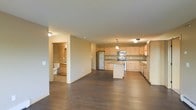 This single-floor space is designed with an open-concept layout that seamlessly connects the living area to the kitchen, creating a spacious and functional environment. The kitchen is a standout feature, showcasing light natural wood cabinetry that extends along two walls, providing generous storage. Stainless steel appliances, including a refrigerator, microwave, and range, add a modern touch, while a central island offers additional counter space and serves as a natural focal point. A pendant light hangs above the island, contributing warmth to the area alongside flush-mount ceiling fixtures throughout. Rich, dark hardwood flooring spans the entirety of the open living space, creating a strong visual contrast against the light-toned cabinetry and neutral beige walls. The open floor plan allows for flexible furniture arrangement in the living area, with clear sightlines across the space. A doorway visible from the main area leads to a bathroom, which features tile flooring and standard fixtures. The overall ambiance is clean, contemporary, and practical, with a neutral palette that provides a versatile backdrop for various interior styles.