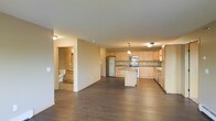 This single-floor space is designed with an open-concept layout that seamlessly connects the living area to the kitchen, creating a spacious and functional environment. The kitchen is a standout feature, showcasing light natural wood cabinetry that extends along two walls, providing generous storage. Stainless steel appliances, including a refrigerator, microwave, and range, add a modern touch, while a central island offers additional counter space and serves as a natural focal point. A pendant light hangs above the island, contributing warmth to the area alongside flush-mount ceiling fixtures throughout. Rich, dark hardwood flooring spans the entirety of the open living space, creating a strong visual contrast against the light-toned cabinetry and neutral beige walls. The open floor plan allows for flexible furniture arrangement in the living area, with clear sightlines across the space. A doorway visible from the main area leads to a bathroom, which features tile flooring and standard fixtures. The overall ambiance is clean, contemporary, and practical, with a neutral palette that provides a versatile backdrop for various interior styles.