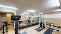 This fitness room offers a functional and practical space designed to support a well-rounded workout routine. The room is outfitted with a selection of exercise equipment, including a Lifestyler treadmill for cardiovascular training and a multi-station cable weight machine for strength and resistance exercises. The light-toned flooring is complemented by black rubber matting beneath the equipment, providing stability and protection during use. Full-length mirrors line one wall, visually expanding the space while allowing users to monitor their form during workouts. The textured ceiling features rows of fluorescent lighting that illuminate the room evenly, ensuring a bright and practical environment at any time of day. The walls carry a neutral, warm beige tone that contributes to a clean and focused atmosphere. Overall, the fitness room is straightforward in its design, prioritizing utility and functionality. It provides residents or guests with convenient access to essential workout equipment within a well-lit, organized, and purpose-built environment.