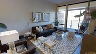 This single-floor space showcases a thoughtfully designed interior that blends modern aesthetics with comfortable living. The living area serves as the centerpiece, featuring a plush dark charcoal sofa adorned with an assortment of decorative throw pillows in neutral and earthy tones. A round glass-top coffee table anchors the seating arrangement, complemented by white upholstered ottomans and side tables that add both functionality and visual contrast. A patterned area rug in soft grey and ivory tones defines the living space and adds warmth to the hardwood flooring beneath. Sleek adjustable floor lamps with metallic gold accents provide stylish ambient lighting, while two framed artwork pieces hang symmetrically on the wall, adding a curated, gallery-like touch. Large floor-to-ceiling windows and a sliding glass door flood the room with natural light and open directly onto a private balcony, offering views of the surrounding exterior. The neutral wall palette in soft white and greige tones creates a calm, cohesive backdrop throughout. The overall ambiance is contemporary and inviting, striking a balance between refined design and everyday comfort.