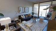 This single-floor space showcases a thoughtfully designed interior that blends modern aesthetics with comfortable living. The living area serves as the centerpiece, featuring a plush dark charcoal sofa adorned with an assortment of decorative throw pillows in neutral and earthy tones. A round glass-top coffee table anchors the seating arrangement, complemented by white upholstered ottomans and side tables that add both functionality and visual contrast. A patterned area rug in soft grey and ivory tones defines the living space and adds warmth to the hardwood flooring beneath. Sleek adjustable floor lamps with metallic gold accents provide stylish ambient lighting, while two framed artwork pieces hang symmetrically on the wall, adding a curated, gallery-like touch. Large floor-to-ceiling windows and a sliding glass door flood the room with natural light and open directly onto a private balcony, offering views of the surrounding exterior. The neutral wall palette in soft white and greige tones creates a calm, cohesive backdrop throughout. The overall ambiance is contemporary and inviting, striking a balance between refined design and everyday comfort.