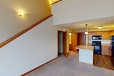 Located at 1910 Hawks Ridge Dr in Madison, Wisconsin, this two-story home offers a well-organized layout spread across two thoughtfully proportioned floors. The main level presents an open-concept living area with soft carpeting and warm-toned trim throughout, flowing seamlessly into a functional kitchen. The kitchen is outfitted with honey-toned wood cabinetry, black appliances including a range, built-in microwave, dishwasher, and refrigerator, complemented by a central island with a light countertop and pendant lighting overhead. Hardwood-style flooring anchors the kitchen space, creating a clean contrast to the carpeted living area. A staircase with wood railings rises from the main level, leading to the upper floor. The upper level features a spacious room with vaulted ceilings, plush carpeting, and a sliding glass door with vertical blinds that allows for natural light and outdoor access. Two full bathrooms are present across both floors, each finished with clean white fixtures and well-lit vanities. Two closets provide practical storage throughout. Wall sconces and ceiling fixtures provide warm, even lighting throughout the home, contributing to an inviting and comfortable ambiance.