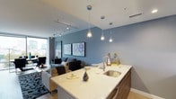 This contemporary single-floor residence showcases a thoughtfully designed open-concept layout that seamlessly connects the kitchen, living, and dining areas. The kitchen is a standout feature, equipped with high-end stainless steel Bosch appliances, including a gas range and built-in microwave, complemented by sleek wood-grain cabinetry and an expansive quartz waterfall island with an undermount sink. Pendant lighting adds a refined touch overhead. The living and dining area is anchored by floor-to-ceiling windows that flood the space with natural light and frame sweeping city views. Track lighting runs across the ceiling, and cool blue-grey walls lend a modern, cohesive aesthetic throughout. The bedroom is a serene retreat, featuring a dramatic custom mural accent wall with an abstract design, floor-to-ceiling windows with urban views, and a distinctive tray ceiling painted in a deep blue-grey hue. The en-suite bathroom is elegantly appointed with large-format marble-style flooring, dark cabinetry with dual sinks, an illuminated vanity mirror, and a tiled shower-tub combination. A walk-in closet is conveniently accessible from the bathroom. The residence also includes an in-unit laundry area, completing this well-appointed urban home.