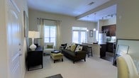 This single-floor space showcases an open-concept layout that seamlessly connects the living area and kitchen, creating a cohesive and functional environment. The living area is anchored by plush, light-toned carpeting and features a dark upholstered sofa accented with patterned throw pillows, complemented by a coordinating accent chair and an olive-toned ottoman. Large windows dressed with floor-length curtains and horizontal blinds flood the space with natural light, enhancing the airy, neutral color palette of soft beige and warm white tones throughout.

The kitchen is defined by dark cabinetry contrasted against lighter countertops and a peninsula with bar-height seating, making it ideal for casual dining or entertaining. Stainless steel appliances, including a full-sized refrigerator, add a modern touch. Pendant lighting hangs above the peninsula, while track lighting illuminates the kitchen workspace. The transition between the carpeted living area and the hard-surface kitchen flooring subtly delineates each zone while maintaining an open feel. Overall, the space conveys a clean, contemporary aesthetic with a comfortable and welcoming ambiance.
