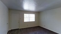 This single-floor space offers a straightforward and functional layout well-suited for comfortable everyday living. The main room is finished with wall-to-wall carpeting that adds warmth underfoot, transitioning to hard flooring near the entryway. A textured popcorn ceiling runs throughout, giving the space a classic residential character. Neutral-toned walls provide a clean, versatile backdrop that accommodates a variety of interior styles and furnishings. A front entry door with a gold-toned handle provides direct access to the room, situated alongside a multi-pane window that allows natural light to filter in, brightening the interior and creating an open, airy feel. The window's gridded design adds a subtle architectural detail to the otherwise understated space. Electrical outlets are conveniently placed along the baseboards, and the overall layout is open and unobstructed, offering flexibility in how the room can be arranged and utilized. The space presents a clean, move-in-ready environment with a simple, practical design that prioritizes usability and ease of living on a single level.