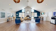 Located at 5613 Crenshaw Rd in Richmond, Virginia, this single-story community clubhouse offers a thoughtfully designed, open-concept layout that serves as a welcoming gathering and administrative hub. The space is defined by dramatic vaulted ceilings that rise to a striking peak, creating an expansive, airy atmosphere throughout. Rich hardwood-style flooring runs continuously across the entire floor, lending warmth and cohesion to the interior.

The main lounge area is a standout feature, anchored by a white brick fireplace framed by a decorative arched mirror and flanked by large windows that flood the space with natural light. Bold teal accent walls provide a vibrant contrast against the crisp white ceiling and trim, while stylish woven pendant light fixtures add a contemporary decorative touch. Comfortable seating arrangements — including a sectional sofa, upholstered armchairs, and a central area rug — create an inviting social environment.

Adjacent to the lounge is a business area equipped with bar-height seating, a wall-mounted television, and a printer station. The entrance corridor features glass-panel doors and private office spaces on either side, outfitted with desks and workstations, ideal for leasing or administrative functions. Abundant windows throughout ensure the entire space remains bright and connected to the outdoors.