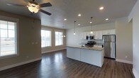 This two-story home presents an open-concept main living area that seamlessly integrates the kitchen and living space into a cohesive, functional layout. Dark hardwood floors run throughout, providing a rich contrast to the neutral beige walls and white cabinetry.

The kitchen serves as a central focal point, anchored by a sizeable island topped with light granite countertops featuring subtle veining. White shaker-style cabinetry lines the walls, offering ample storage, while stainless steel appliances — including a refrigerator, range, and built-in microwave — provide a modern, polished finish. Pendant lights hang above the island, adding targeted task lighting that complements the recessed lighting installed throughout the ceiling.

The adjacent living area benefits from natural light streaming through multiple windows fitted with horizontal blinds. A ceiling fan is positioned overhead, enhancing airflow and comfort. The textured ceiling adds a subtle dimensional quality to the space. Together, the kitchen and living areas create a welcoming, well-lit environment suited for both everyday living and entertaining across the home's two floors.