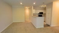 This single-floor space is designed with an open-concept layout that seamlessly connects the living area and kitchen. The living space features neutral beige walls, white baseboards, and soft wall-to-wall carpeting, creating a clean, understated aesthetic. Recessed lighting and a flush-mount ceiling fixture provide even illumination throughout.

The kitchen is defined by a white peninsula counter that doubles as a breakfast bar, offering both prep space and a natural visual divider between the two areas. White cabinetry lines the walls, complemented by a tiled backsplash and stainless steel appliances, including an over-the-range microwave. A track lighting fixture above the kitchen adds focused task lighting. The kitchen also includes a sink and additional counter space along the back wall.

The transition between the kitchen and living area is smooth and unobstructed, making the overall space feel open and functional. The consistent use of white trim, neutral paint, and light carpeting throughout contributes to a cohesive, bright interior. The layout is practical and well-suited for everyday living and entertaining.