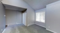 This single-floor residence offers a clean, functional layout with a straightforward design. The bedroom is a standout feature of the space, showcasing vaulted ceilings that lend an airy, open feel to the room. Soft gray carpet covers the floor, adding warmth and comfort underfoot, while light gray walls provide a neutral and versatile backdrop throughout.

A large window fitted with white horizontal blinds allows natural light to filter into the bedroom, creating a bright and welcoming atmosphere. Glimpses of greenery through the window suggest a pleasant outdoor setting. The room is equipped with a built-in closet featuring sliding white doors, offering practical storage space. Baseboard heaters line the lower edges of the walls, ensuring efficient climate control throughout the colder months.

The textured popcorn ceiling adds a classic residential touch, complementing the overall simplicity of the space. Clean white baseboards frame the carpeted floors, contributing to the tidy, well-maintained appearance of the interior. The layout is open and uncluttered, making the room highly adaptable to a variety of furniture arrangements and personal styles. Overall, the space presents a comfortable and functional living environment with practical features throughout.