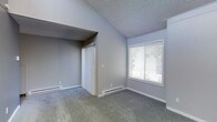 This single-floor residence offers a clean, functional layout with a straightforward design. The bedroom is a standout feature of the space, showcasing vaulted ceilings that lend an airy, open feel to the room. Soft gray carpet covers the floor, adding warmth and comfort underfoot, while light gray walls provide a neutral and versatile backdrop throughout.

A large window fitted with white horizontal blinds allows natural light to filter into the bedroom, creating a bright and welcoming atmosphere. Glimpses of greenery through the window suggest a pleasant outdoor setting. The room is equipped with a built-in closet featuring sliding white doors, offering practical storage space. Baseboard heaters line the lower edges of the walls, ensuring efficient climate control throughout the colder months.

The textured popcorn ceiling adds a classic residential touch, complementing the overall simplicity of the space. Clean white baseboards frame the carpeted floors, contributing to the tidy, well-maintained appearance of the interior. The layout is open and uncluttered, making the room highly adaptable to a variety of furniture arrangements and personal styles. Overall, the space presents a comfortable and functional living environment with practical features throughout.
