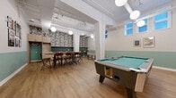Located at 87 Mann Ave in Ottawa's Sandy Hill neighbourhood, this well-appointed amenity room is designed as a multi-functional social and recreational space. The open-plan layout is anchored by warm light wood flooring and a two-tone wall treatment featuring sage green wainscoting paired with crisp white upper walls, giving the space a fresh, contemporary aesthetic. Exposed white structural columns and overhead ductwork lend the room an industrial character, while globe pendant lights provide warm, even illumination throughout. A full-size pool table with a teal felt surface commands the central area, complemented by a foosball table nearby for added recreation. Along one wall, a bar-style counter with wood cabinetry, dark teal subway tile backsplash, and built-in appliances serves as a kitchenette, with high-top tables and wooden chairs arranged for casual dining or socializing. A bold tropical-print feature wall adds a vibrant design accent to this area. Tucked in one corner, a cozy lounge nook features a sleek linear fireplace set into a wood-panelled wall, flanked by built-in bench seating and accompanied by a grey sectional sofa and accent chairs on a concrete floor. Windows along one wall allow natural light to filter in, enhancing the welcoming ambiance of this versatile communal space.