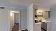 This single-story residence offers a clean, functional layout designed for comfortable everyday living. The interior is finished with dark hardwood-style flooring that runs throughout, providing a warm contrast to the soft gray walls and bright white crown molding that accents each room.

The kitchen is a standout feature, equipped with classic cabinetry in a warm tone, under-cabinet range ventilation, a stainless steel sink, and light-colored countertops. A peninsula with a breakfast bar extends into the adjacent living area, creating an open and connected flow that is ideal for both casual dining and entertaining.

Crown molding details add a refined architectural touch to the ceilings, enhancing the overall sense of polish and care in the design. A hallway leads to the remaining rooms of the residence, which benefit from the same consistent flooring and neutral wall tones, creating a cohesive and calming ambiance throughout. Ample natural and overhead lighting ensures each space feels bright and welcoming. The overall design balances simplicity with practicality, making this a well-appointed single-floor residence suited to a variety of lifestyles.