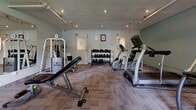 Located at 550 Mornington in London, Ontario, this fitness centre offers a well-organized and functional workout space across a single floor. The room features soft teal-toned walls, a textured concrete ceiling with recessed pot lights and exposed pipe-mounted track lighting, and durable modular carpet tile flooring in neutral tones that provides comfort underfoot during exercise.

The space is thoughtfully equipped with a varied selection of fitness equipment to support a full-body workout routine. Cardio offerings include a treadmill, elliptical trainer, and stationary bike, while strength training is supported by a multi-function cable machine, a Life Fitness adjustable weight bench, and a dedicated free weight rack stocked with a range of dumbbells. A large wall-mounted mirror along one side of the room enhances the sense of openness and allows users to monitor their form during exercise.

Natural light filters in through a window on one wall, complemented by the recessed ceiling lighting to keep the space bright and welcoming. Two interior doors provide access to adjoining areas, and a wall clock and exercise reference posters are mounted near the entrance for added convenience. A small potted plant adds a touch of warmth to the otherwise functional environment. This fitness centre provides residents with a convenient and comprehensive on-site amenity for their daily fitness needs.