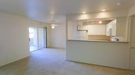 This single-floor residence offers a clean, open-concept layout designed for comfortable everyday living. The spacious living area is finished with soft wall-to-wall carpeting and features a ceiling fan that contributes to a relaxed, airy ambiance. A sliding glass door along one wall invites natural light into the space and provides access to an outdoor area, enhancing the sense of openness throughout.

At the heart of the home, the kitchen is defined by a raised pass-through counter that connects it seamlessly to the living area. White cabinetry lines the walls, complemented by recessed and track lighting overhead that keeps the space well-lit and functional. Built-in appliances, including a microwave and refrigerator, are neatly integrated into the cabinetry, contributing to a cohesive and efficient kitchen design.

The overall aesthetic throughout is light and neutral, with consistent warm-toned walls and ceilings that create a welcoming and harmonious feel. The thoughtful open floor plan encourages a natural flow between living and kitchen spaces, making the home feel connected and versatile. The single-floor layout adds convenience and accessibility, making it a practical and inviting space overall.
