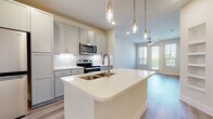 This single-floor residence presents a clean, contemporary design throughout its well-organized layout. The open-concept kitchen serves as the centerpiece of the living space, showcasing gray shaker-style cabinetry extending to the ceiling for ample storage. White quartz countertops span generously across the perimeter and a large central island, which features an undermount sink with a brushed nickel faucet and seating overhang. A full suite of stainless steel appliances includes a French-door refrigerator, electric range, and over-the-range microwave. Subway tile backsplash and under-cabinet lighting add polished finishing touches, while pendant lighting with gold accents illuminates the island below. Light wood-look flooring flows through the kitchen and living areas, which connect seamlessly and benefit from natural light through a sliding glass door leading to a balcony. The bedroom is finished with plush neutral carpet and a window offering outdoor views. The bathroom features a gray vanity with white quartz countertop and a framed mirror. A dedicated laundry closet houses a stacked washer and dryer unit, adding everyday convenience to this thoughtfully designed space.