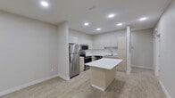 Located at 921 S Taylor Rd in McAllen, Texas, this single-story space showcases a clean, modern aesthetic throughout. The open-concept kitchen serves as the centerpiece, featuring stainless steel appliances including a full-size refrigerator, electric range, and built-in microwave. Flat-panel cabinetry in white and warm beige tones lines the walls, complemented by bright white countertops and a single-basin sink. A freestanding center island provides additional workspace and a natural gathering point within the kitchen. Recessed lighting illuminates the space uniformly, enhancing the contemporary feel.

Light wood-look flooring runs consistently throughout, lending warmth and cohesion to each area. The bedroom is a calm, well-proportioned room outfitted with a ceiling fan, recessed lighting, and a window dressed with layered roller shades that allow for adjustable natural light. Neutral gray walls contribute to a clean, understated atmosphere.

The bathroom is finished with the same light flooring and features a modern vanity with white countertop, a standard toilet, and a sliding glass-enclosed walk-in shower. The overall design is minimalist and contemporary, creating a cohesive, move-in-ready environment.