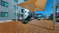 Located in Maricopa, Arizona, this outdoor recreational space is thoughtfully designed to serve residents of the surrounding multi-story community. The play area centers on a robust, multi-component playground structure featuring green slides, climbing elements, and activity panels, all set atop a generous wood chip safety surface that cushions the entire play zone. Large shade sails are strategically positioned overhead, providing essential relief from the Arizona sun and making the space comfortable for extended outdoor play.

The perimeter is framed by desert-adapted landscaping, incorporating decorative gravel beds, low-water shrubs, and young trees that blend seamlessly with the arid Sonoran climate. Tall street lamps are distributed throughout, ensuring the area remains well-lit into the evening hours. A distinct open zone within the wood chip area offers additional unstructured play space for younger children.

Adjacent to the playground, a covered pergola-style structure shelters picnic tables, creating a communal gathering spot for families. Swaying palm trees line the transitional path toward a nearby pool and lounge area, complete with shaded seating and vibrant umbrellas. The space is cleanly bordered by concrete sidewalks and is fully integrated with the surrounding residential buildings, making it a convenient and inviting outdoor amenity for the entire community.