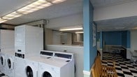 This single-floor laundry facility is designed with practicality and convenience in mind. The space features a row of modern front-load washing machines and dryers, providing ample capacity for users. Overhead fluorescent lighting ensures the area is well-lit throughout, creating a bright and functional environment. A textured ceiling adds a subtle architectural detail, while a distinctive blue structural column provides a pop of color against the otherwise neutral white and grey tones of the space. The layout is thoughtfully organized, with machines arranged for easy access and efficient workflow. A partial dividing wall separates sections of the room, lending a sense of structure without compromising the open feel of the space. Wooden seating is available, offering a comfortable spot for users to wait. Informational signage is posted on the wall, providing guidance for users of the facility. The clean, well-maintained surfaces and orderly arrangement of equipment reflect a commitment to upkeep and usability. Overall, this laundry facility delivers a straightforward, no-frills environment that prioritizes functionality and ease of use for its patrons.